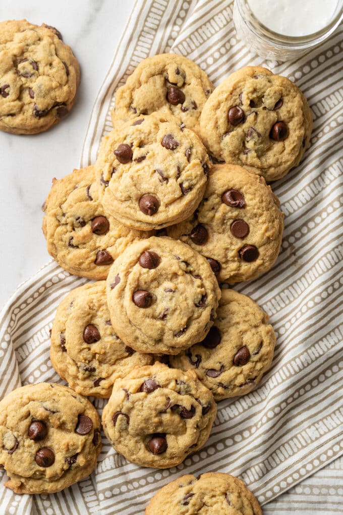 Pile of soft chocolate chip cookies on top of a napkin, with a glass of milk to the side.