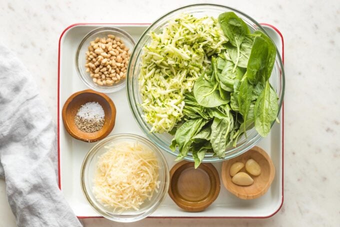 Prep bowls containing grated zucchini, spinach and basil leaves, Parmesan cheese, garlic, pine nuts, olive oil, salt and pepper.