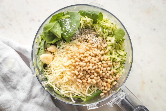 Grated zucchini, spinach and basil, and seasonings in the bowl of a food processor, not yet blended.