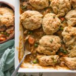 Overhead view of a large casserole dish filled with a homemade chicken pot pie topped with savory Parmesan biscuit crumbles, with one spoonful removed to a nearby wooden bowl.
