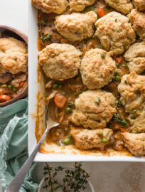 Overhead view of a large casserole dish filled with a homemade chicken pot pie topped with savory Parmesan biscuit crumbles, with one spoonful removed to a nearby wooden bowl.