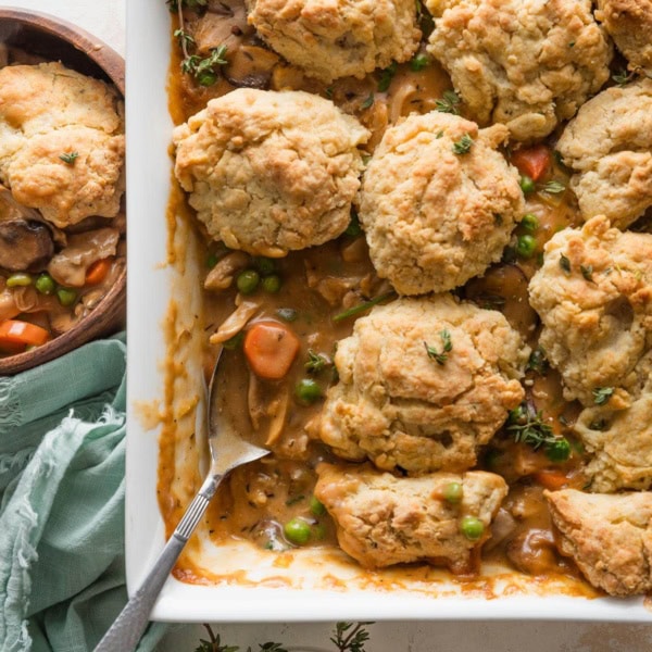 Overhead view of a large casserole dish filled with a homemade chicken pot pie topped with savory Parmesan biscuit crumbles, with one spoonful removed to a nearby wooden bowl.