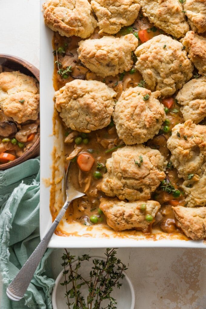 Overhead view of a large casserole dish filled with a homemade chicken pot pie topped with savory Parmesan biscuit crumbles, with one spoonful removed to a nearby wooden bowl.
