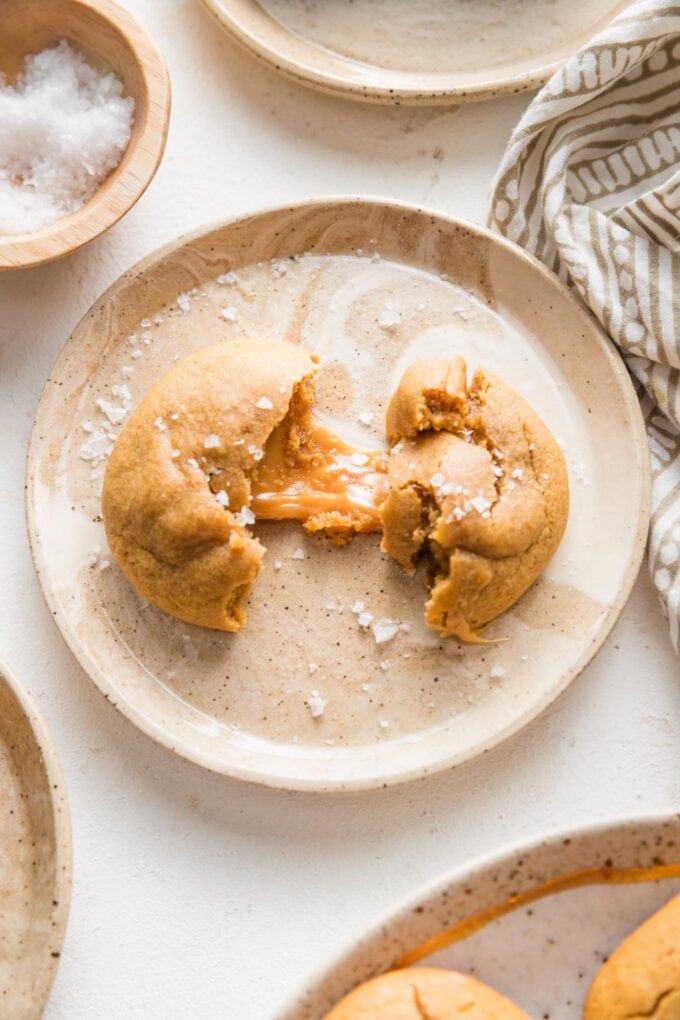 Small brown plate with a sea salt-sprinkled caramel molasses cookie on it, half pulled apart to reveal a chewy caramel inside.