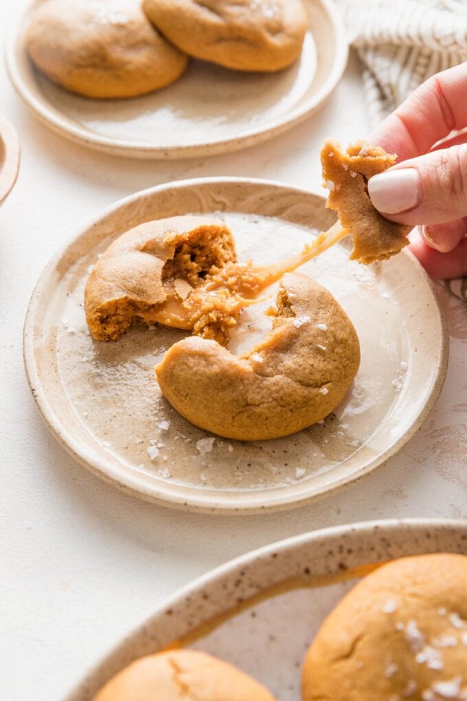 A caramel molasses cookie sitting on a small plate, with a woman's hand pulling part of the cookie and a tender caramel off screen so the caramel stretches out.