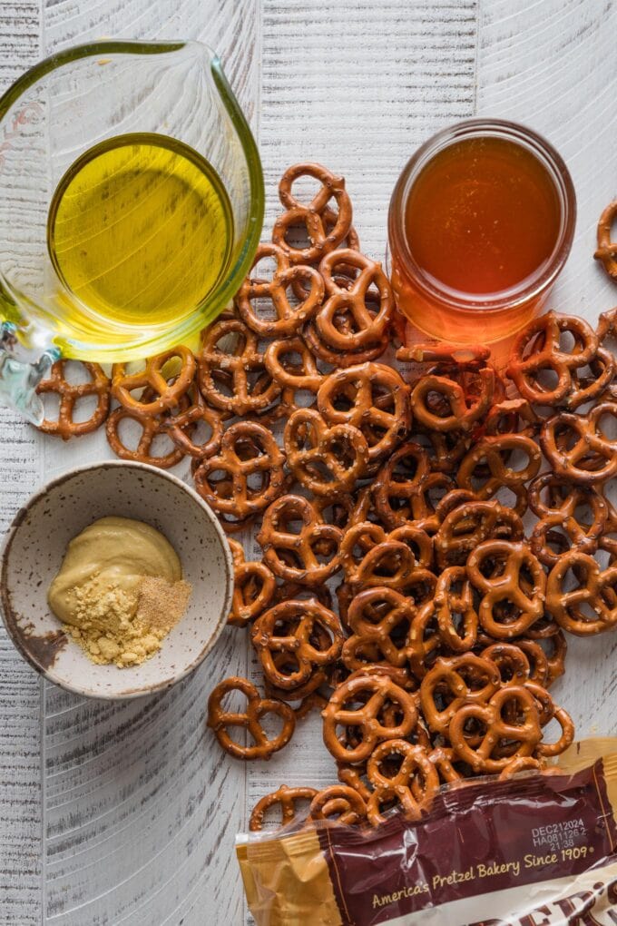 Overhead view of a wooden countertop scattered with a bag of mini pretzels, olive oil, honey, whole grain mustard, dried mustard, and garlic powder, ready to mix into a homemade seasoning blend.