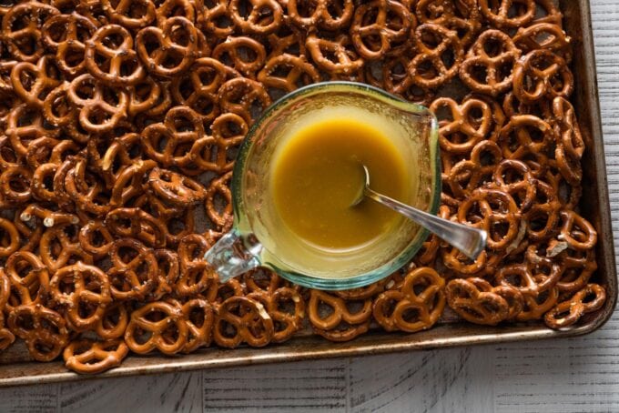 Landscape image of a large baking sheet spread with miniature pretzels and a small glass liquid measuring cup with a honey mustard seasoning blend mixed together in the middle, ready to pour over.