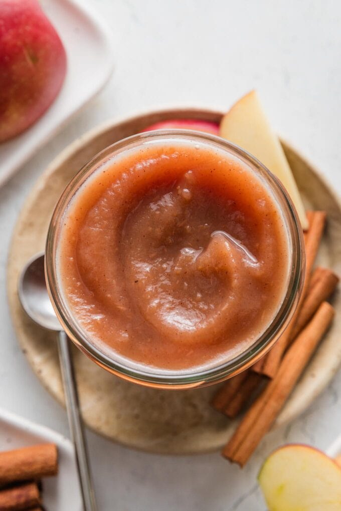 Small jar full of homemade Instant Pot apple butter resting on a plate with apple slices and cinnamon sticks.