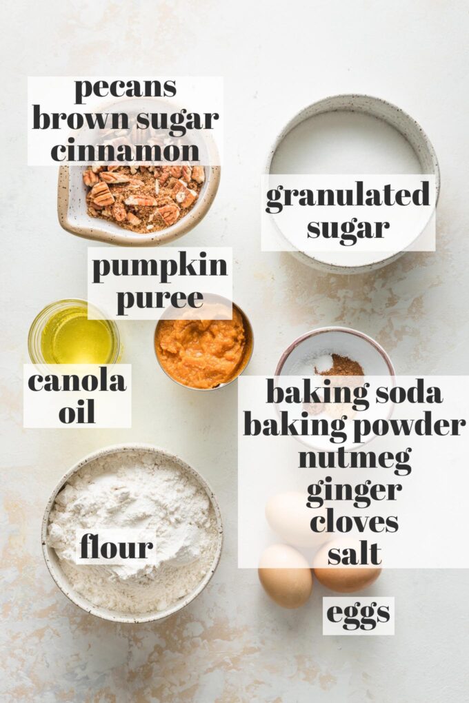 Labeled overhead photo of flour, white sugar, brown sugar, ground cinnamon, chopped pecans, canola oil, pumpkin puree, eggs, nutmeg, ginger, cloves, baking soda, and baking powder all measured into prep bowls and ready to bake.