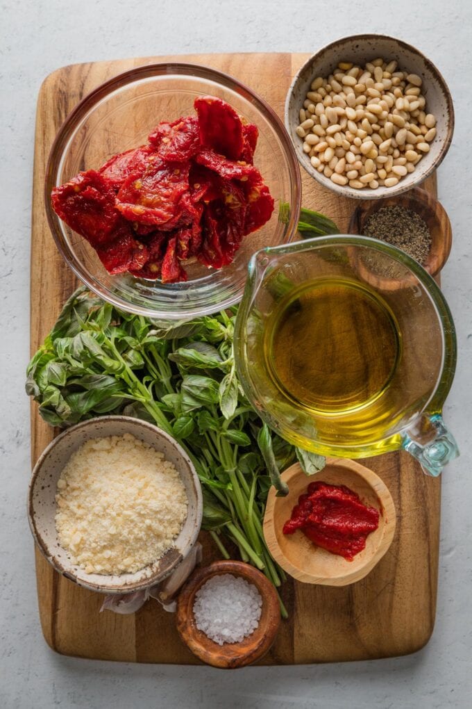 Cutting board set with the ingredients for homemade red pesto: sun-dried tomatoes, fresh basil, pine nuts, olive oil, Parmesan, tomato paste, salt and pepper.