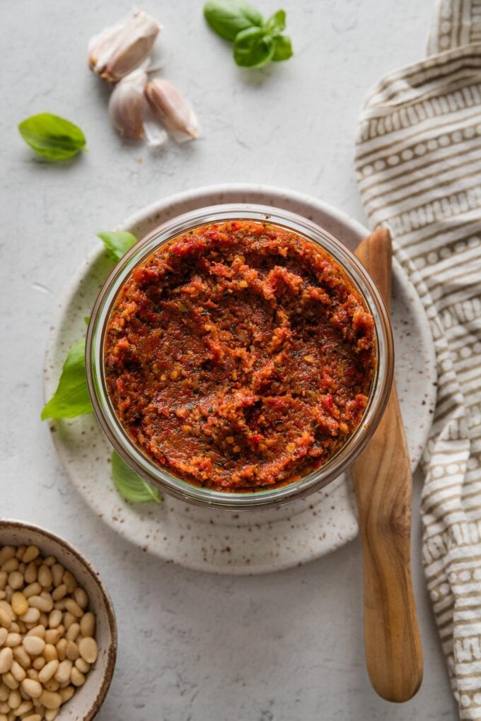 Countertop set with fresh basil leaves, garlic cloves, pine nuts, and a jar of homemade sun-dried tomato pesto.