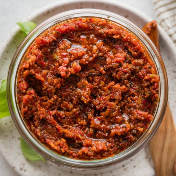Homemade pesto rosso (sun-dried tomato pesto) stored in a glass jar with a wooden serving spreader on a plate nearby.