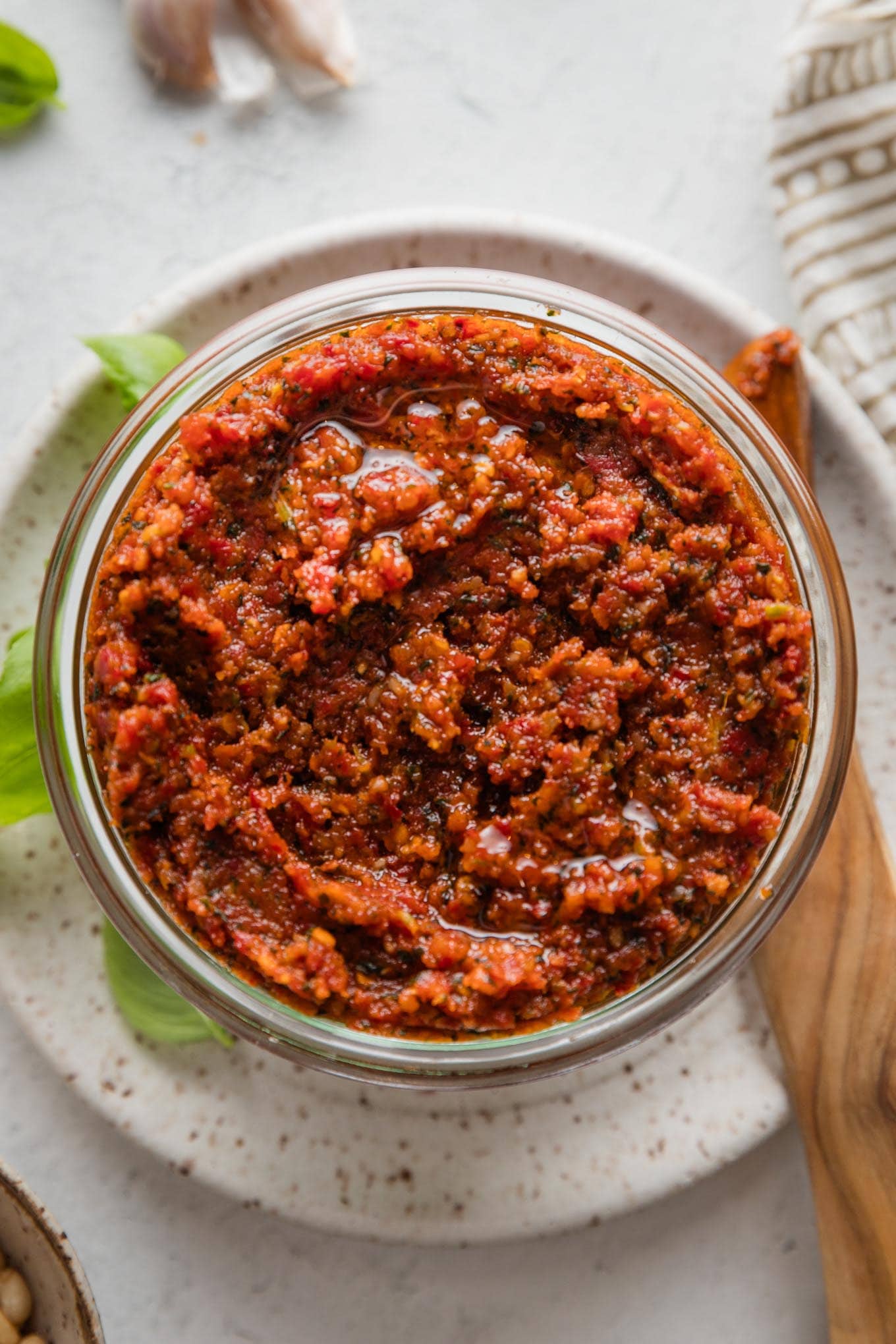 Homemade pesto rosso (sun-dried tomato pesto) stored in a glass jar with a wooden serving spreader on a plate nearby.