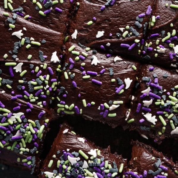 Close up overhead view of a blackout chocolate buttermilk sheet cake topped with ghost and bat-themed Halloween sprinkles and sliced into small squares.