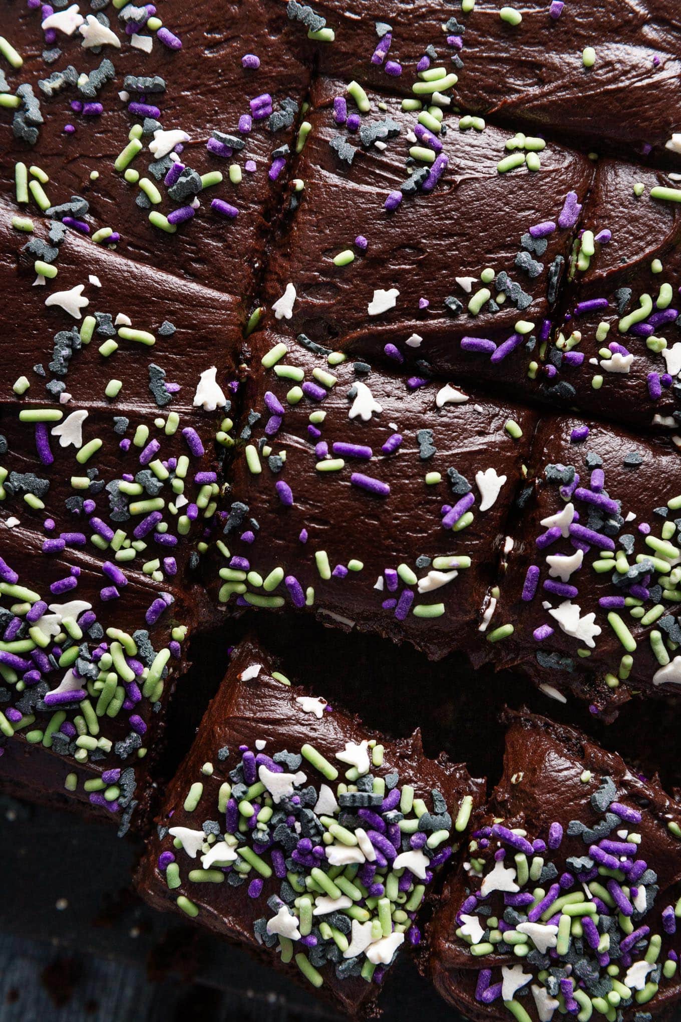 Close up overhead view of a blackout chocolate buttermilk sheet cake topped with ghost and bat-themed Halloween sprinkles and sliced into small squares.