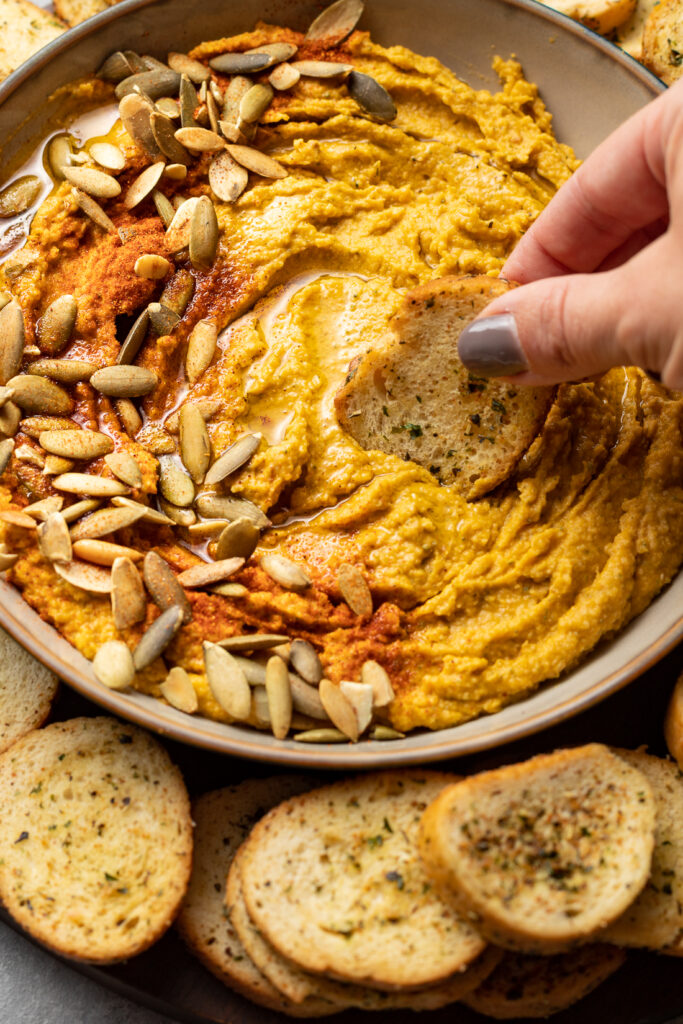 Hand dipping a garlic toast into a bowl of pumpkin hummus.