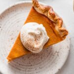 Close up view of a slice of bourbon molasses pumpkin pie cut and served on a small dessert plate with whipped topping and a light dusting of ground cinnamon on top.