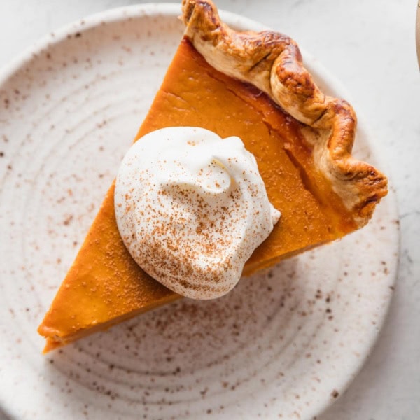 Close up view of a slice of bourbon molasses pumpkin pie cut and served on a small dessert plate with whipped topping and a light dusting of ground cinnamon on top.