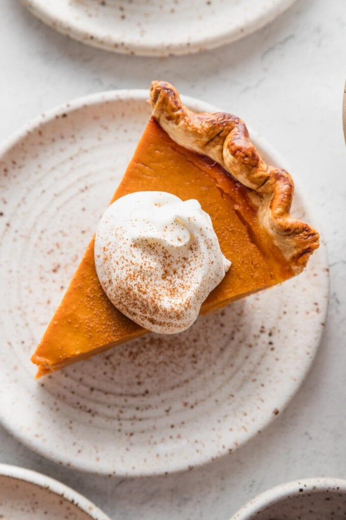 Close up view of a slice of bourbon molasses pumpkin pie cut and served on a small dessert plate with whipped topping and a light dusting of ground cinnamon on top.