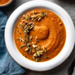 Bowl of creamy carrot red pepper soup with bread, thyme, and pumpkin seeds.