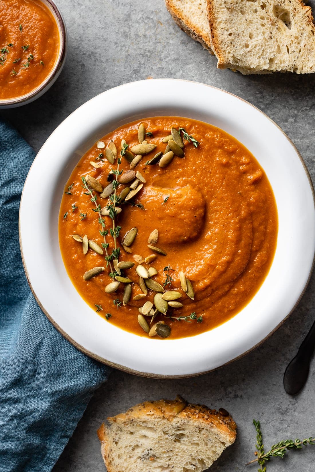 Bowl of creamy carrot red pepper soup with bread, thyme, and pumpkin seeds.
