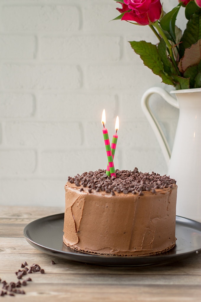 A tiny chocolate cake with mocha buttercream frosting and chocolate chips, with candles lit for a birthday.