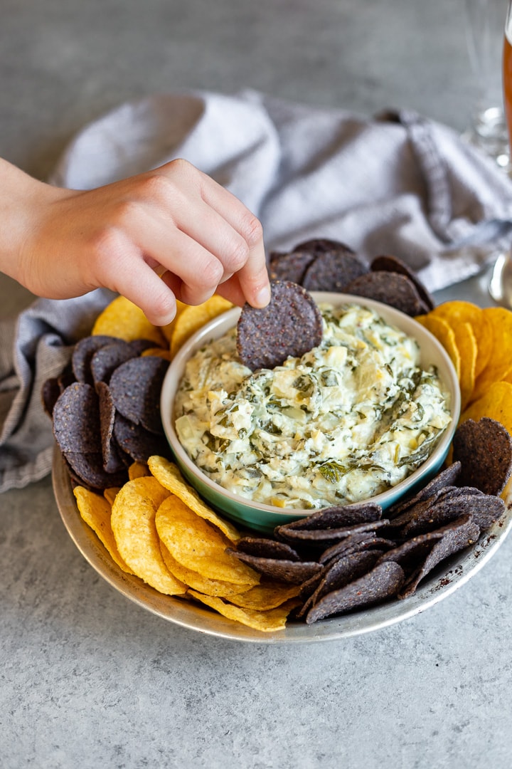 A hand dipping a blue tortilla chip into a bowl of piping hot spinach artichoke dip.