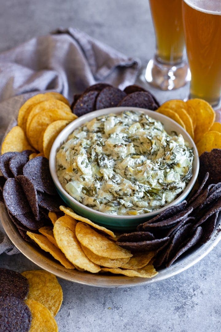 A bowl of skinny crockpot spinach artichoke dip served with tortilla chips and cold beer.