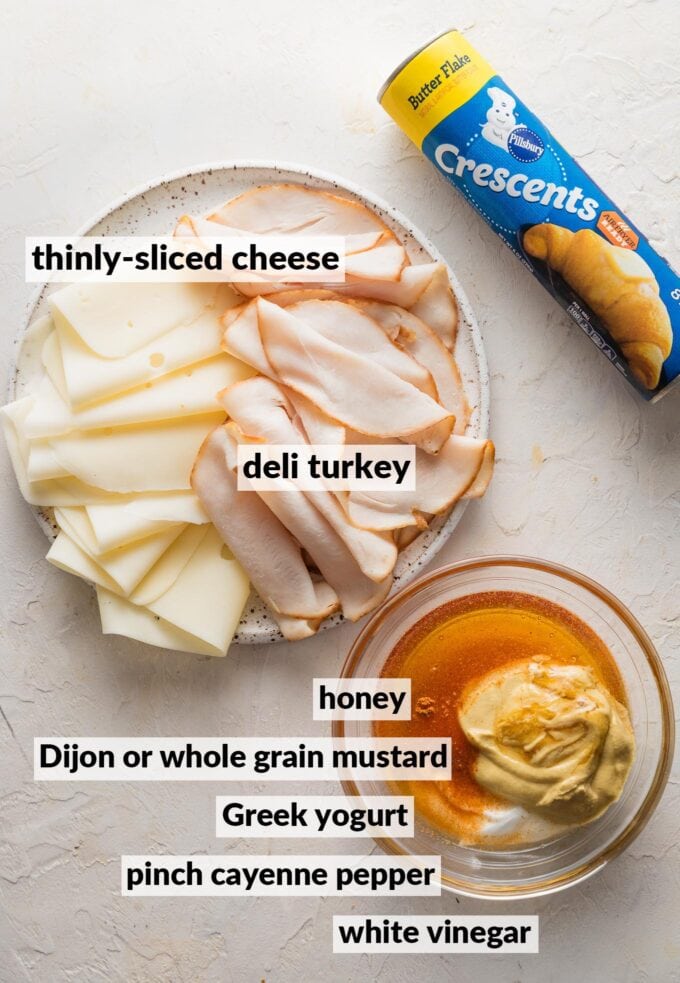 Labeled overhead photo of a tube of crescent roll dough, a plate full of thinly-sliced deli turkey and cheese, and bowl with honey, Greek yogurt, Dijon mustard, and a smidge of white vinegar and cayenne pepper.