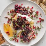 Platter with a wedge of baked brie topped with a cranberry pomegranate sauce, toasted pecans, honey, and fresh rosemary sprigs.