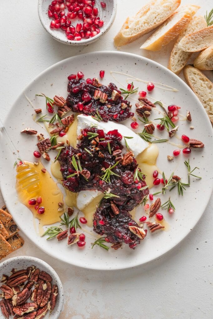 Platter with a wedge of baked brie topped with a cranberry pomegranate sauce, toasted pecans, honey, and fresh rosemary sprigs.