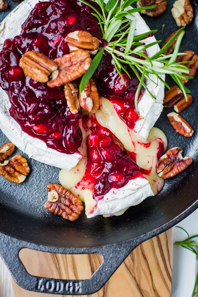 A large wheel of brie baked in a cast-iron skillet, topped with cranberry pomegranate sauce, pecans, and fresh rosemary, with a wedge partially removed.