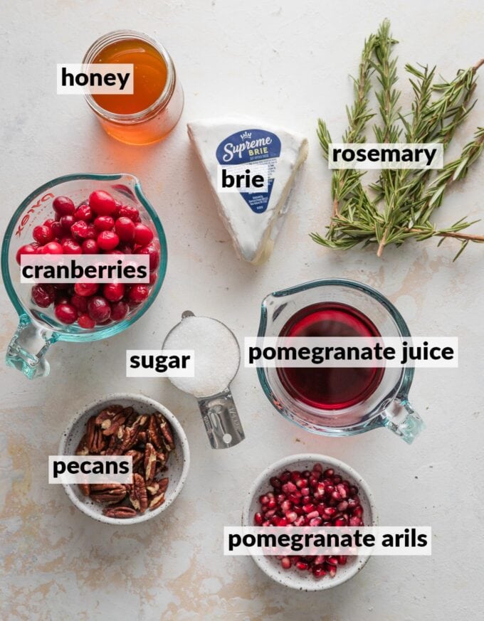 Labeled overhead photo of fresh cranberries, pomegranate juice and pomegranate arils, a wedge of brie, honey, pecans, sugar, and fresh rosemary sprigs.