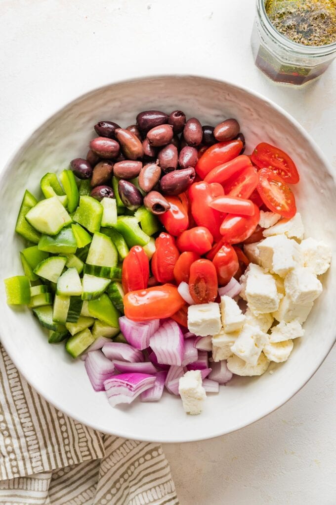 White bowl with cucumber, bell pepper, cherry tomatoes, Kalamata olives, feta cheese, and red onion inside.