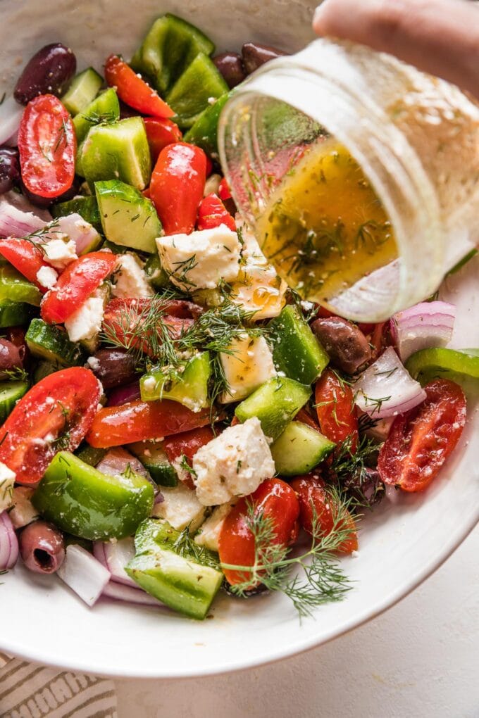 Close up image of salad dressing being poured from a small jam jar over a homemade Greek salad.