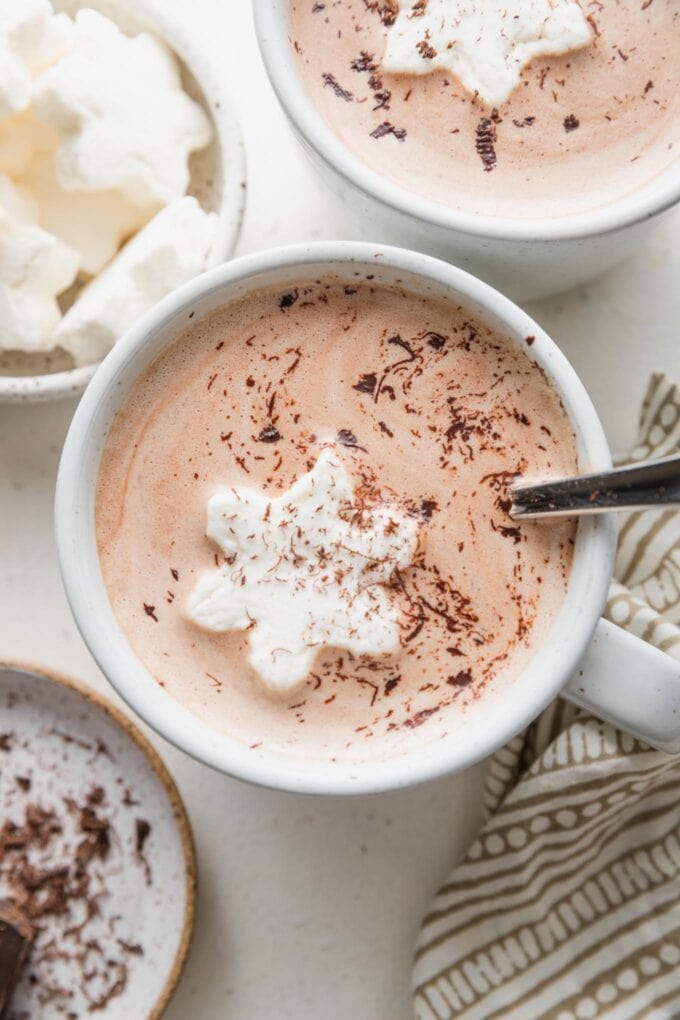 Close up overhead view of two mugs of homemade hot cocoa, from a chocolate cream base, served with snowflake-shaped marshmallows and chocolate shavings.