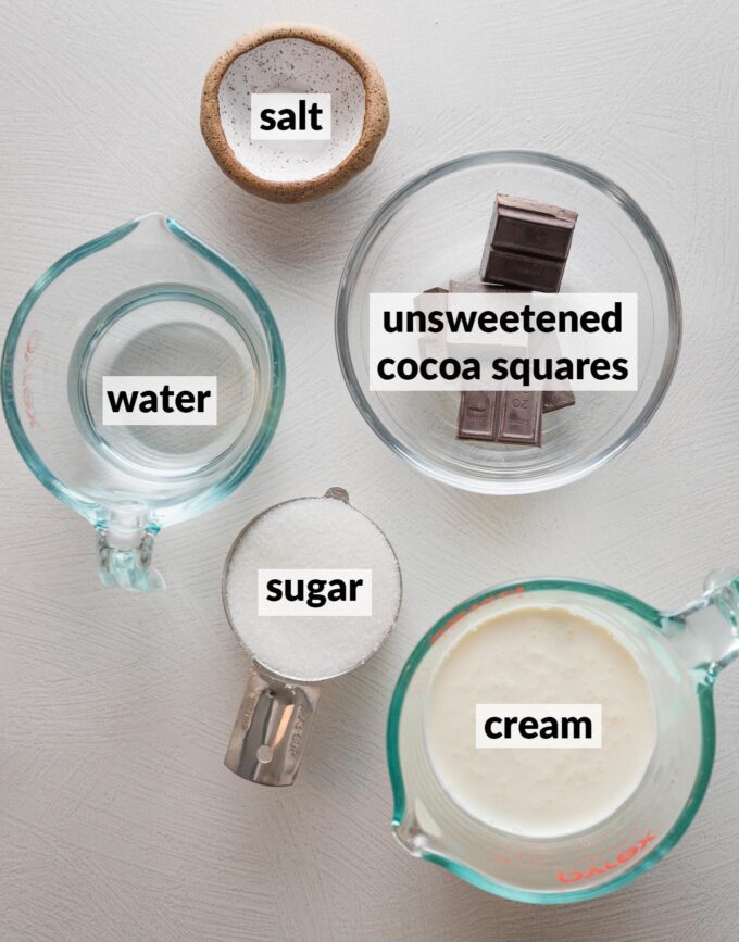 Overhead view of ingredients for a homemade cream hot cocoa mix: unsweetened cocoa squares, cream, sugar, water, and salt.