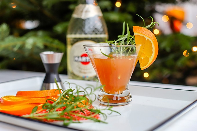 A short champagne flute filled with a light pink orange pomegranate Prosecco cocktail, garnished with rosemary and an orange slice.