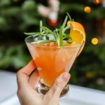 A hand holding up an orange pomegranate Prosecco cocktail in front of a Christmas tree.