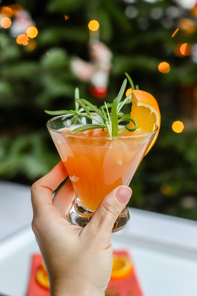 A hand holding up an orange pomegranate Prosecco cocktail in front of a Christmas tree.