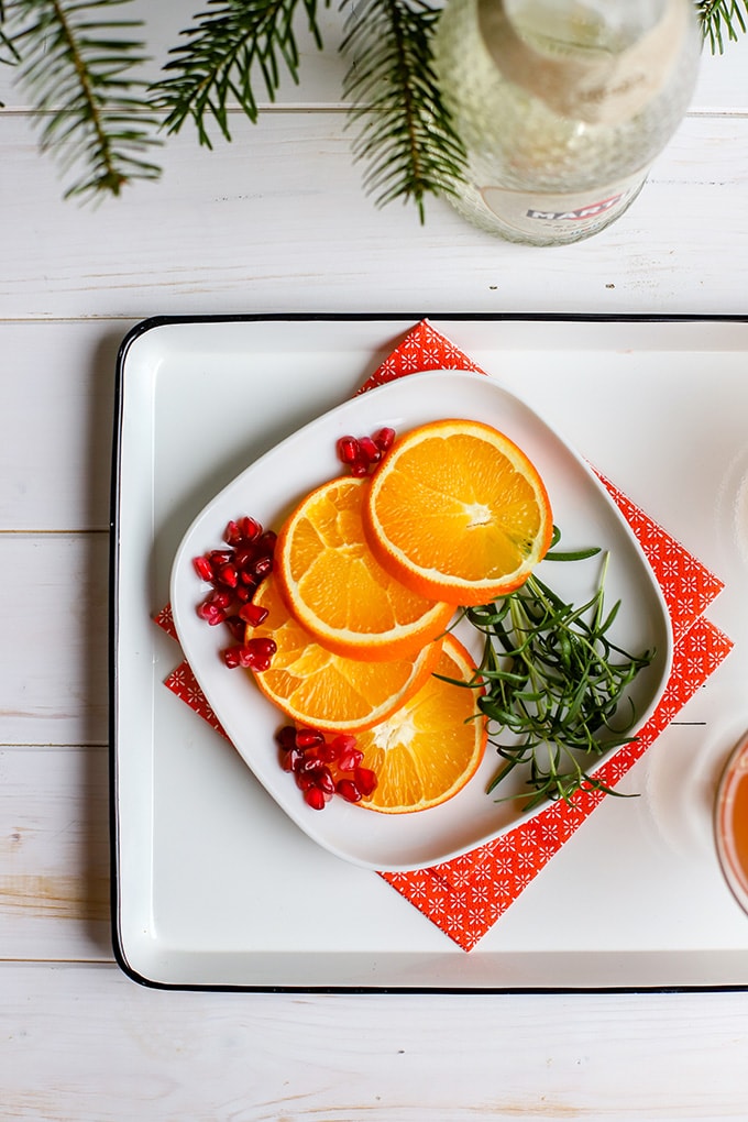 A small square white plate holding garnishes for orange pomegranate Prosecco cocktails.