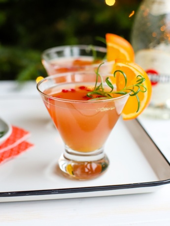 A short champagne flute filled with a light pink orange pomegranate Prosecco cocktail, garnished with rosemary and an orange slice.