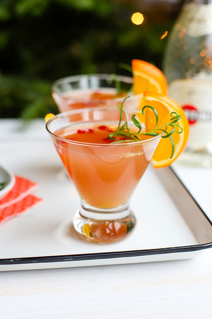 A short champagne flute filled with a light pink orange pomegranate Prosecco cocktail, garnished with rosemary and an orange slice.