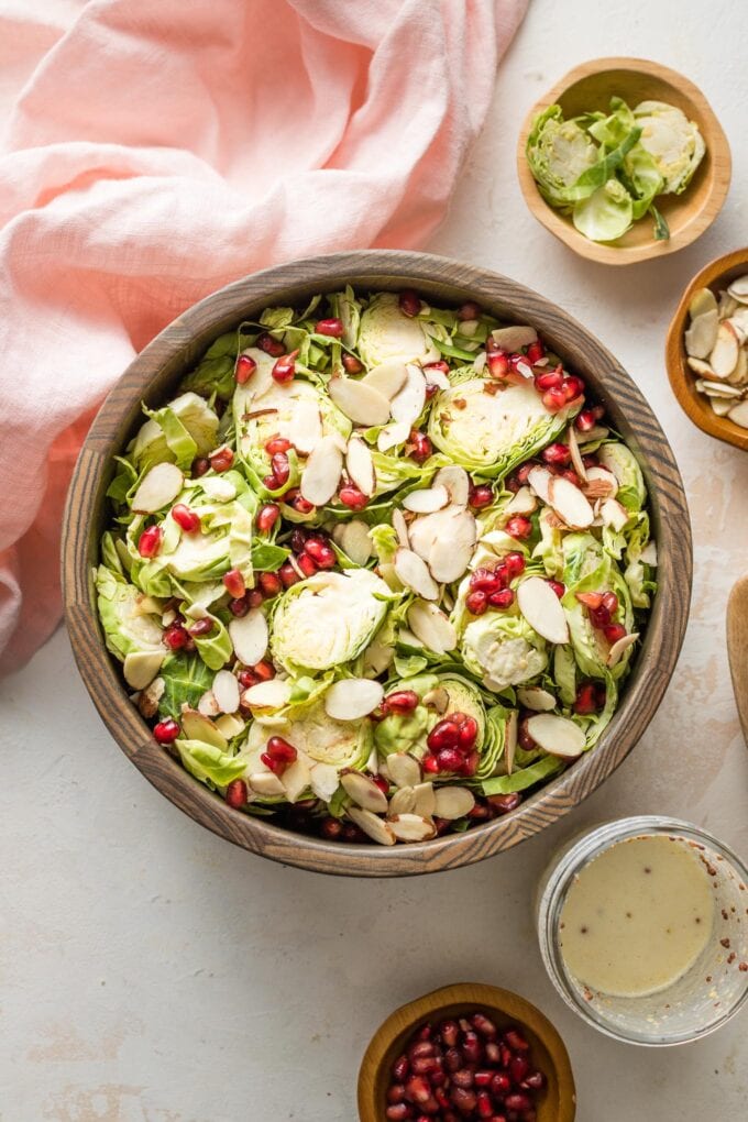 Wooden bowl holding a salad with Brussels sprouts, pomegranate seeds, toasted almonds, and a tangy buttermilk dressing.