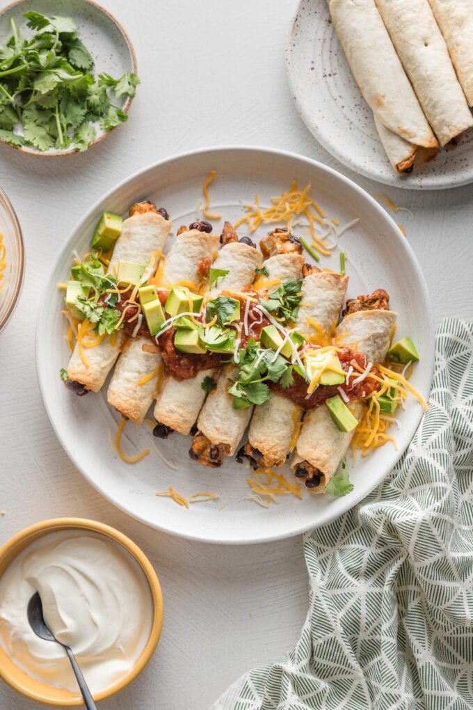 Countertop set with a large plate of chicken black bean taquitos surrounded by fresh cilantro, sour cream, and extra garnishes.