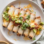 White plate filled with oven-baked chicken and black bean taquitos, dressed with extra cheese, diced avocado, and fresh cilantro leaves.