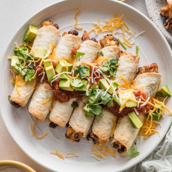 White plate filled with oven-baked chicken and black bean taquitos, dressed with extra cheese, diced avocado, and fresh cilantro leaves.