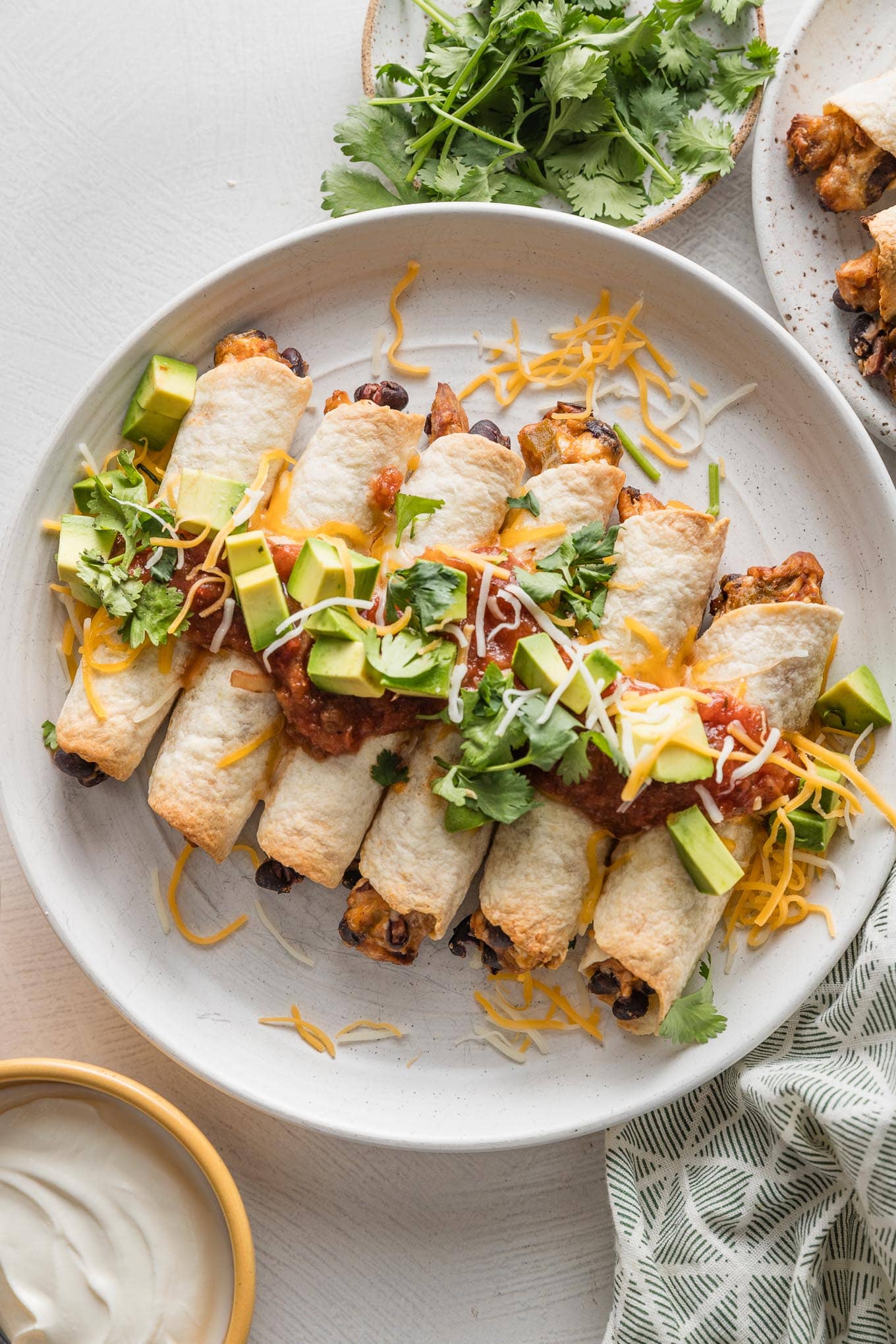 White plate filled with oven-baked chicken and black bean taquitos, dressed with extra cheese, diced avocado, and fresh cilantro leaves.