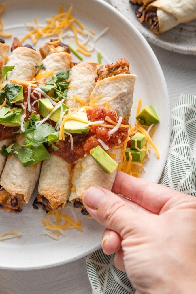 Hand holding up a chicken black bean taquito straight out of the oven and placed on a serving plate.