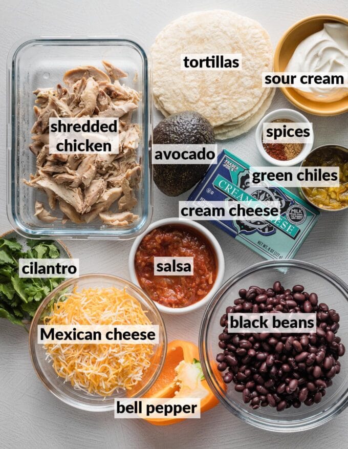 Labeled overhead photo of shredded chicken, black beans, sour cream, tortillas, avocado, spices, canned green chiles, salsa, cream cheese, fresh cilantro, bell pepper, and shredded Mexican cheese, all on the counter and ready to make into taquitos.